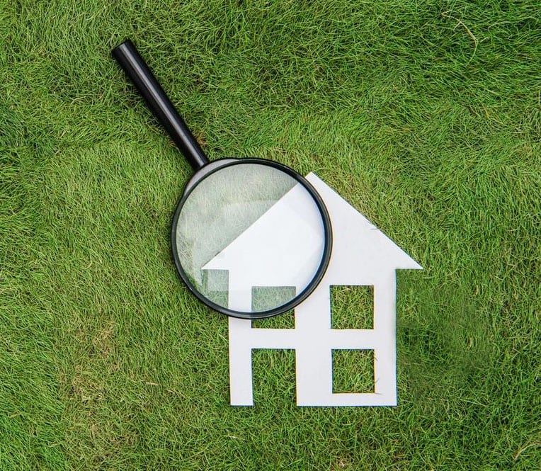 A magnifying glass is on the grass next to a paper cut out of a house.